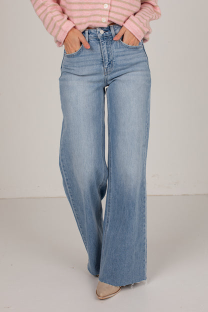 A person wearing light-wash blue wide-leg jeans, a pink striped long-sleeve top, and beige pointed-toe flats, standing against a plain off-white studio background.
