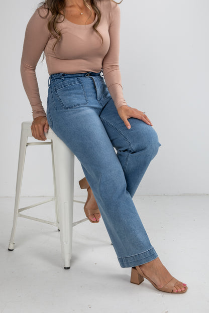 Woman wearing blue jeans and a beige top sitting on a white stool against a white background