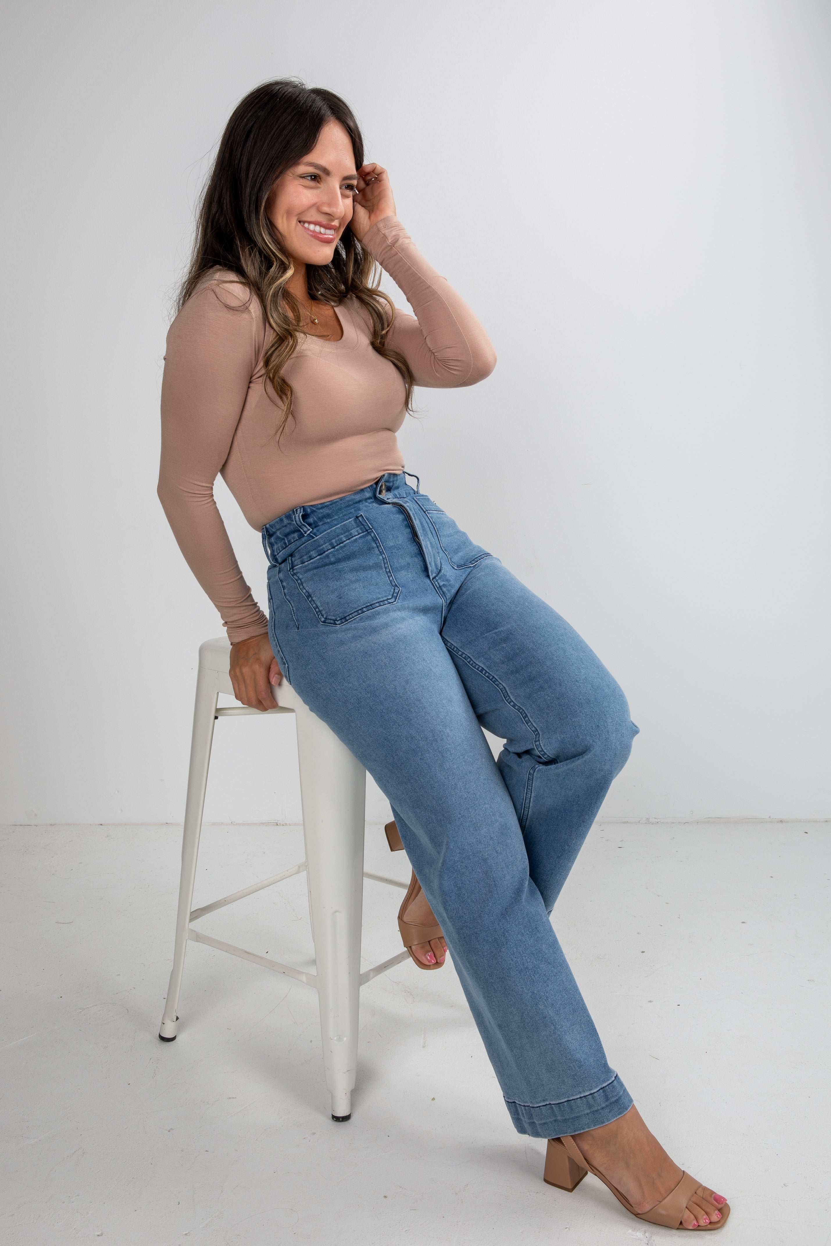 Woman wearing a beige top and blue jeans sitting on a white stool against a white background