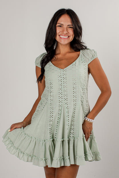 A woman modeling a light sage green eyelet mini dress with short puff sleeves, a V-neckline, and a tiered ruffled hem against a plain light gray studio background.