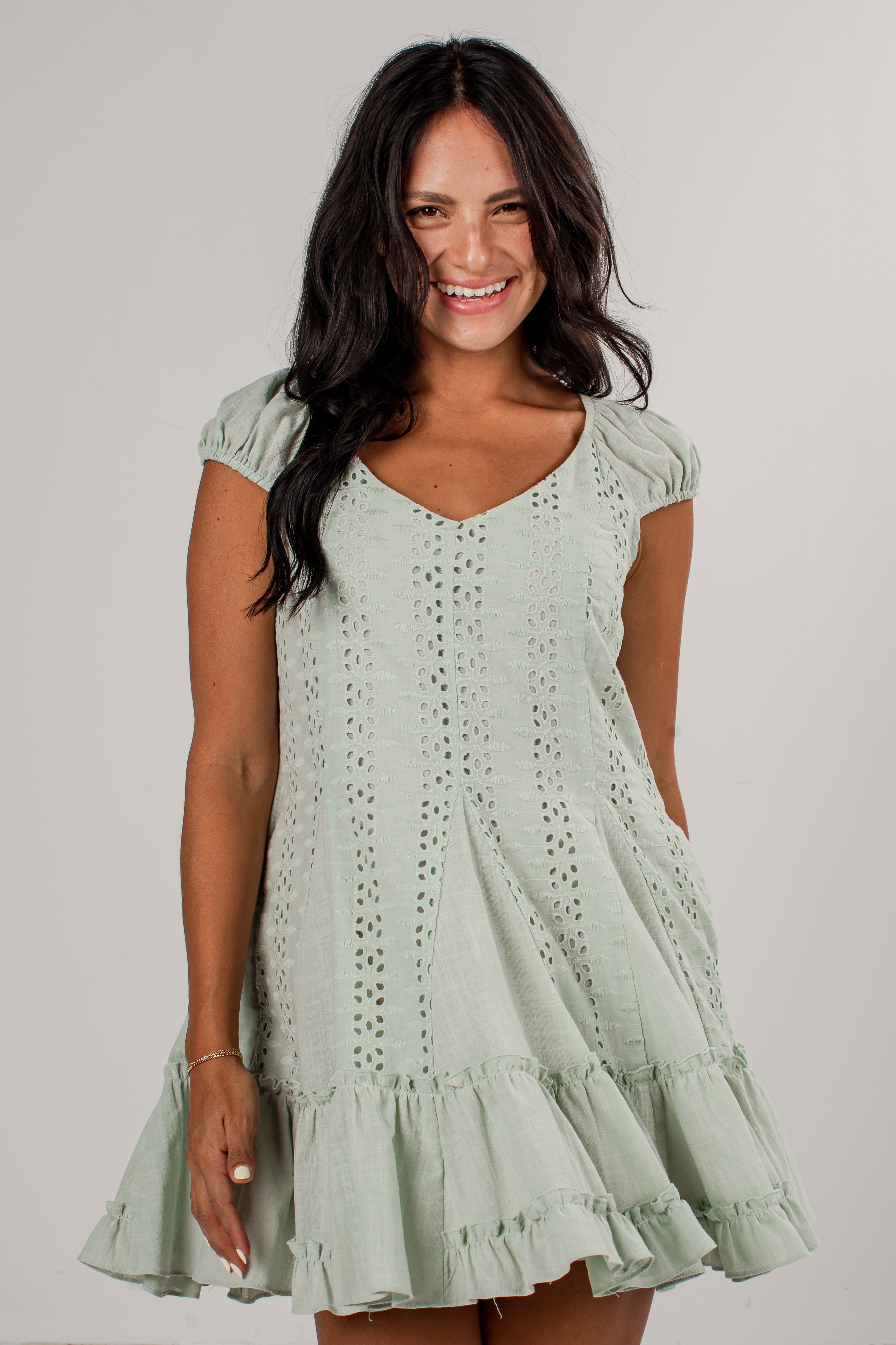 A woman wearing a light sage green eyelet mini dress with cap sleeves and a ruffled hem, standing against a plain light gray studio background.