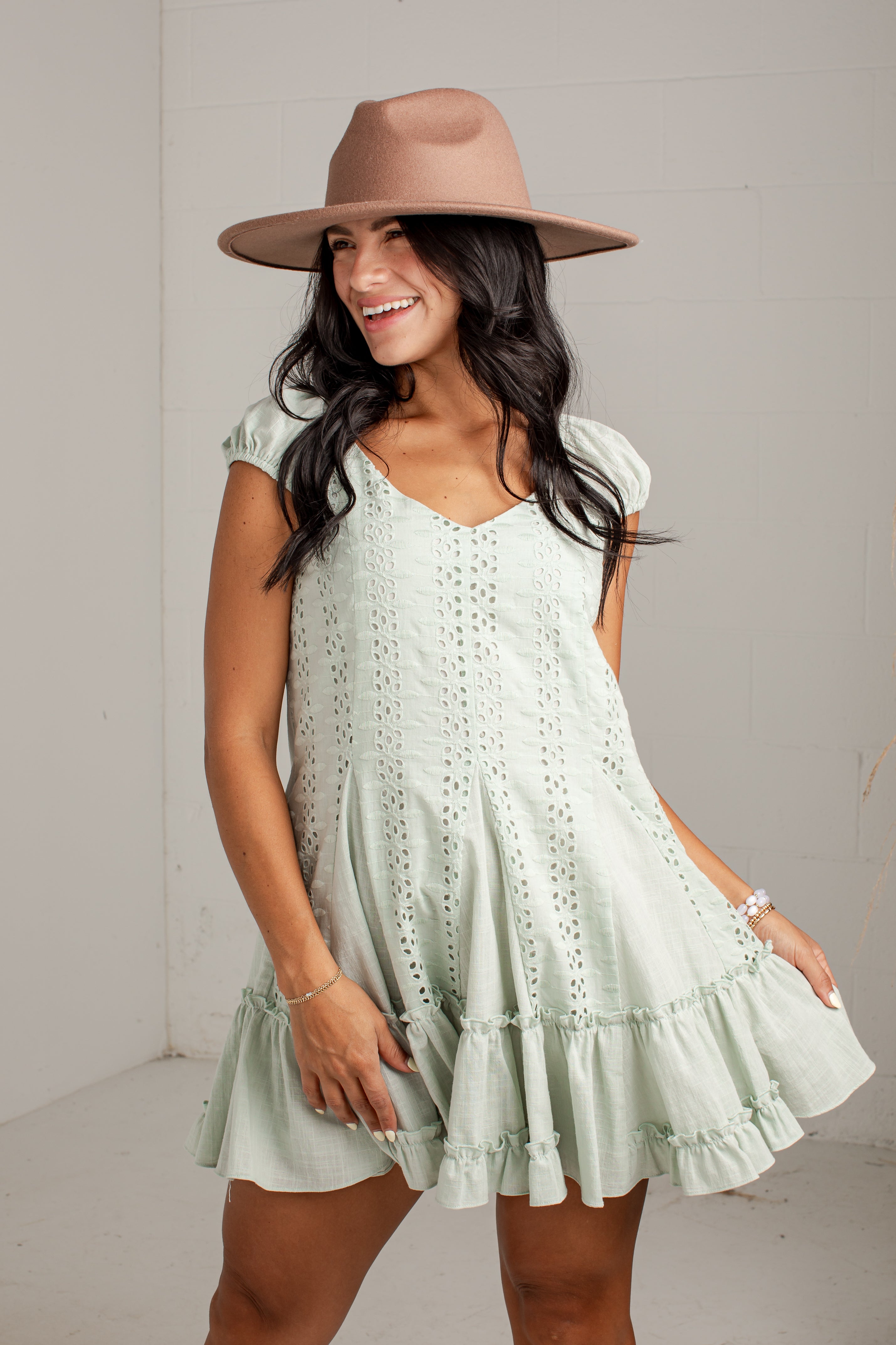 A woman wearing a light sage green eyelet mini dress with tiered ruffles and a matching wide-brim tan hat, posing against a light gray wall.