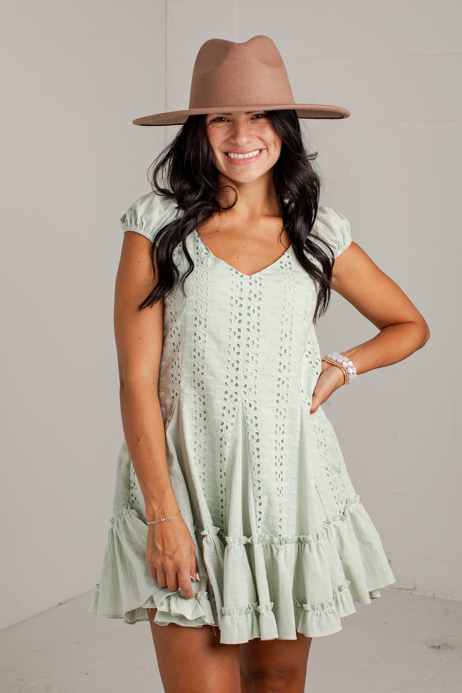 A woman wearing a light sage green eyelet mini dress with ruffled tiers and a brown wide-brim fedora hat, posing against a light gray wall.