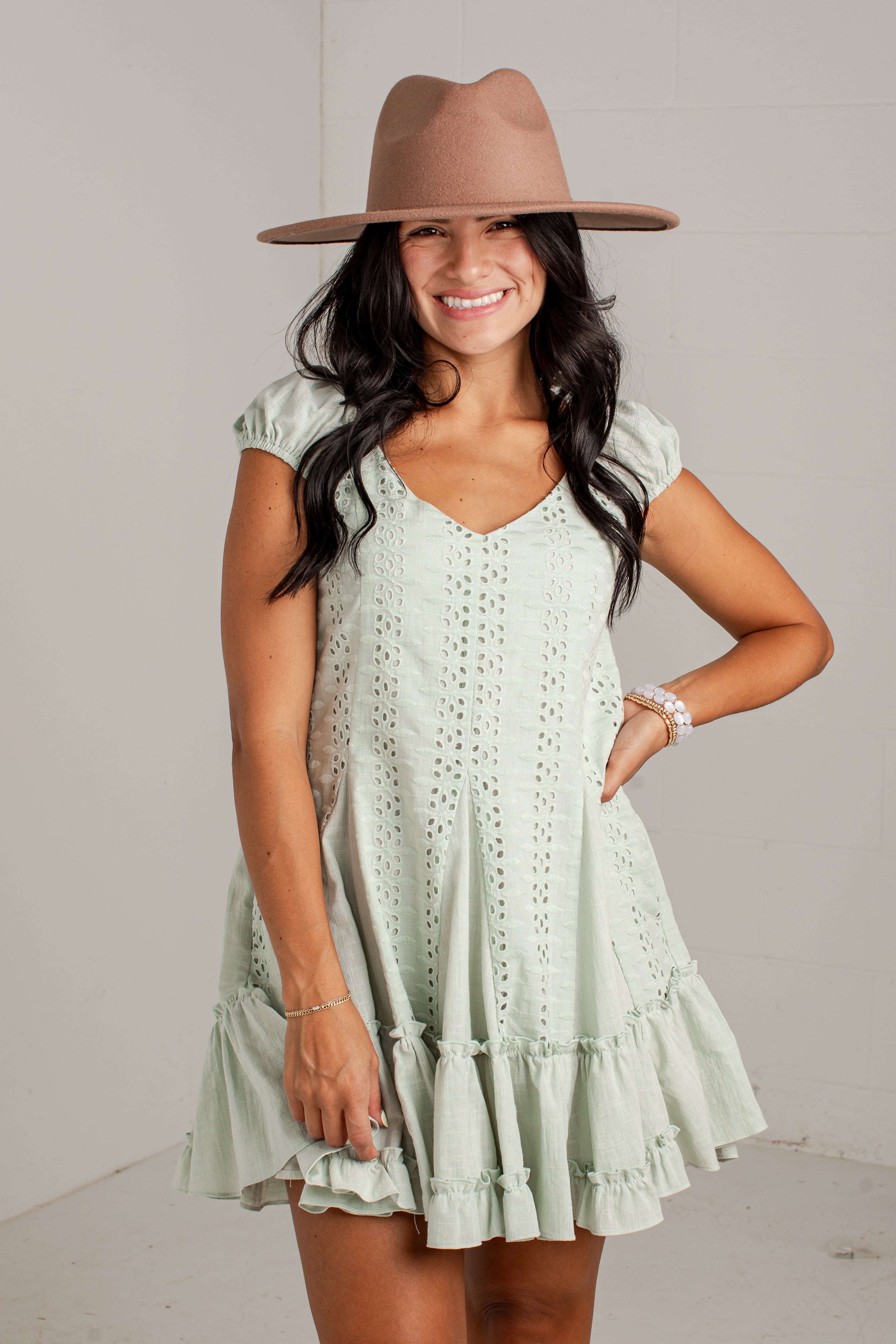 A woman wearing a light sage green eyelet mini dress with ruffled tiers and a brown wide-brim fedora hat, posing against a light gray wall.