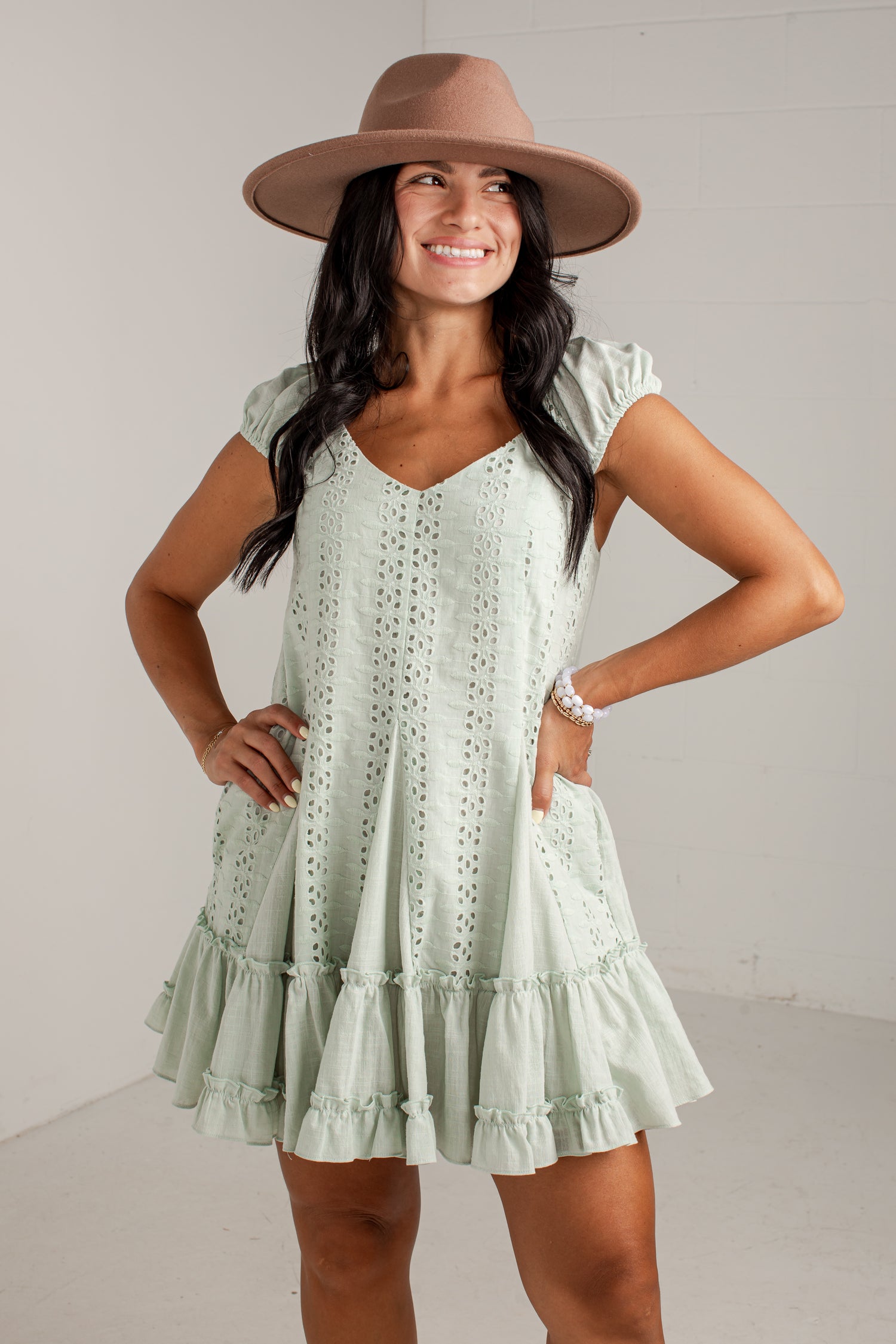 A woman modeling a light sage green eyelet mini dress with a tiered ruffle hem, styled with a wide-brimmed brown hat against a white studio background.