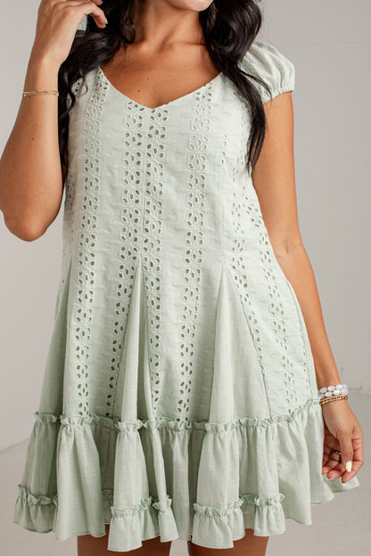 A woman wearing a pale mint green eyelet mini dress with short puff sleeves, a V-neckline, and tiered ruffle hems, shown against a plain light gray studio background.