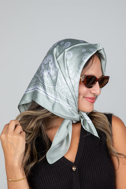 Woman wearing a light green headscarf with white patterns and sunglasses on a gray background