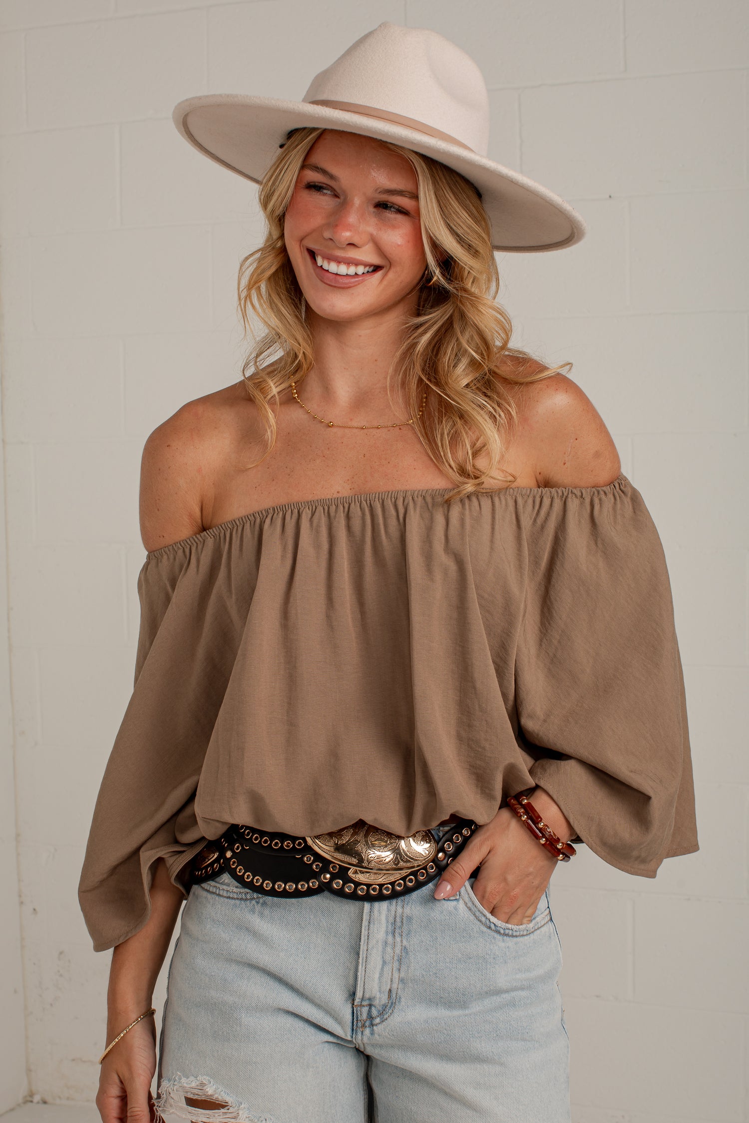 A woman wearing a taupe brown off-the-shoulder puff-sleeve top, a cream wide-brim hat, and light-wash distressed jeans, standing against a white brick wall.