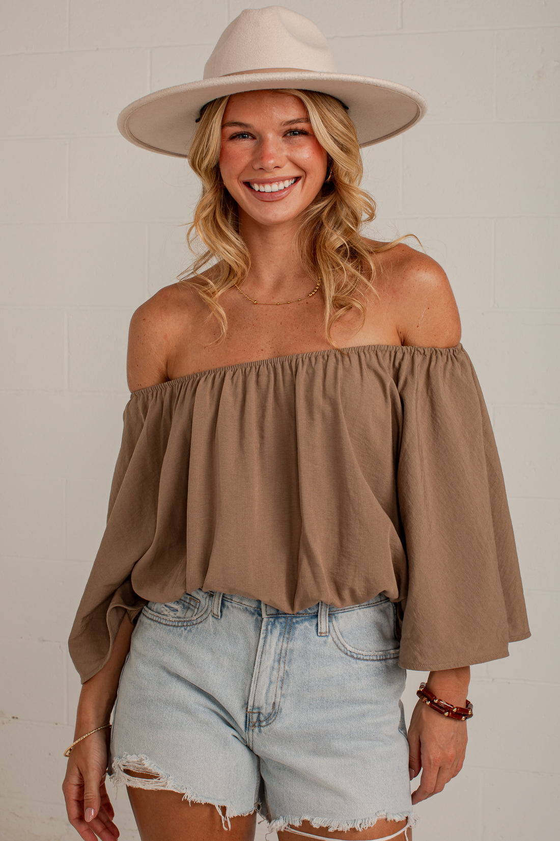 A woman wearing a taupe brown off-the-shoulder long-sleeve top, light-wash denim shorts, and a beige wide-brim hat, posing against a light gray brick wall.
