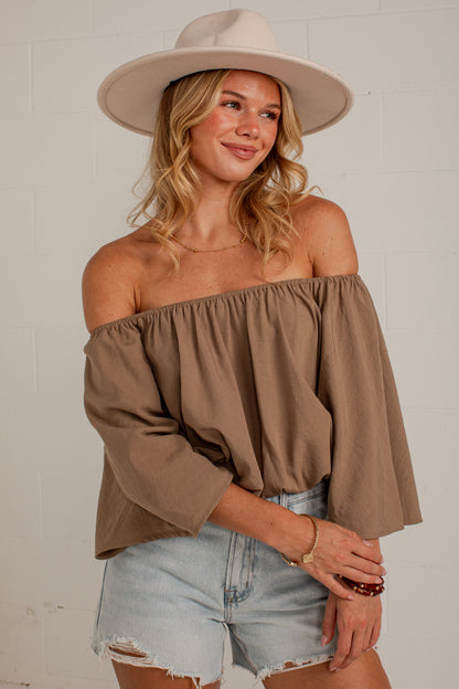 A woman wearing a taupe brown off-the-shoulder top with three-quarter sleeves, paired with light-wash denim shorts and a beige wide-brim hat, posing against a white brick wall.