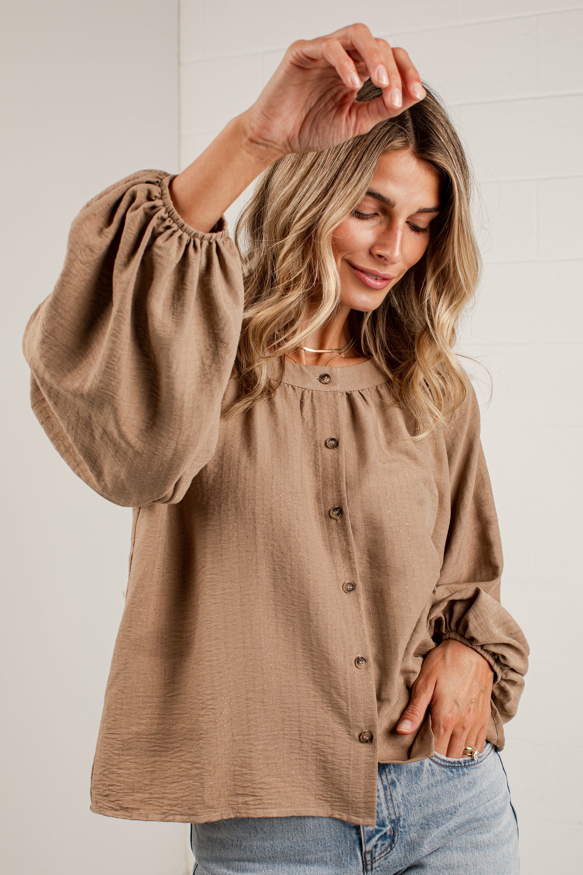A woman wearing a brown long-sleeve blouse with gathered puff sleeves and a button-down front, paired with light blue jeans, posing against a white paneled wall.