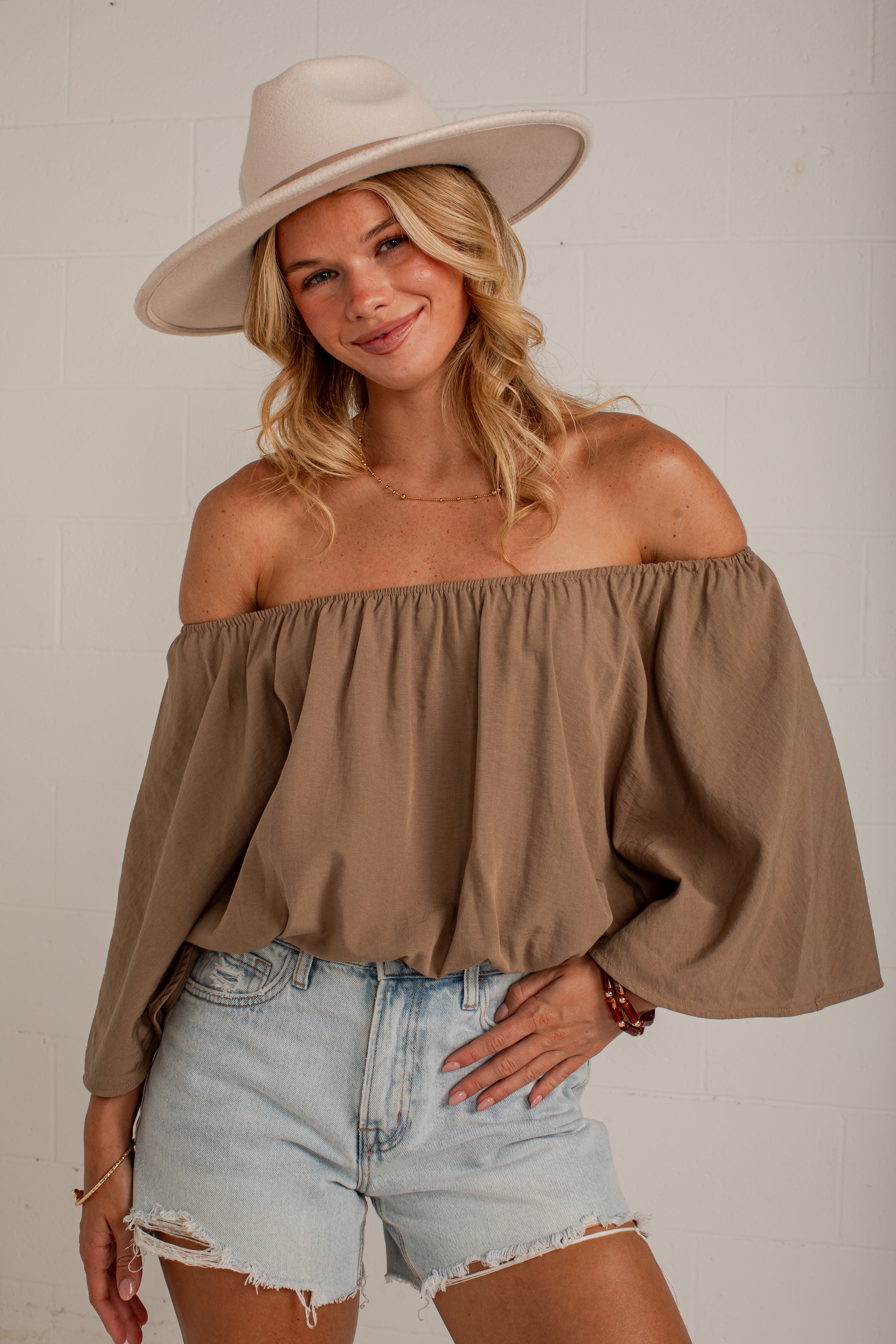 A woman wearing a taupe-brown off-the-shoulder long-sleeve top, light-wash denim shorts, and a beige wide-brim hat, posing against a white brick wall.
