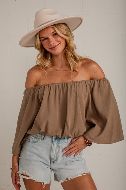 A woman wearing a taupe-brown off-the-shoulder long-sleeve top, light-wash denim shorts, and a beige wide-brim hat, posing against a white brick wall.