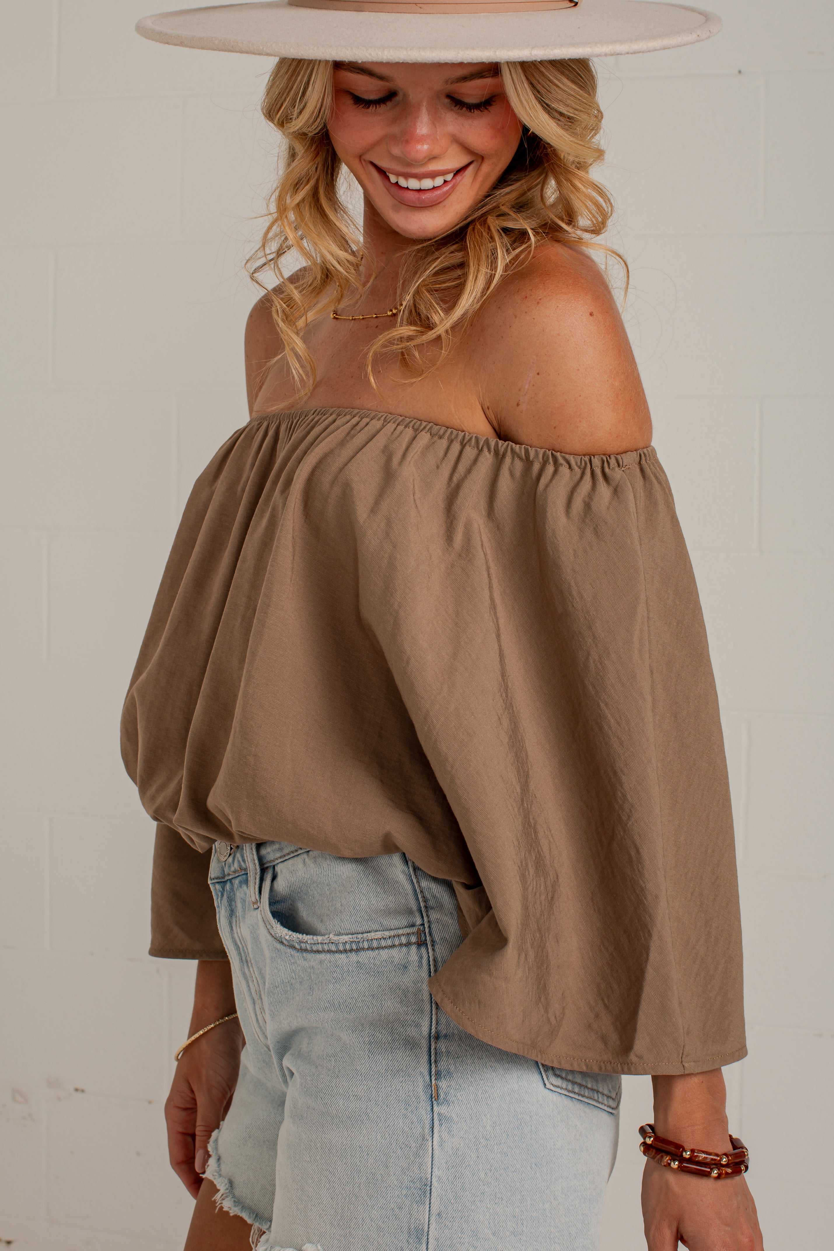 A woman wearing a brown off-the-shoulder balloon-sleeve top and light-wash denim shorts, standing against a white brick wall.