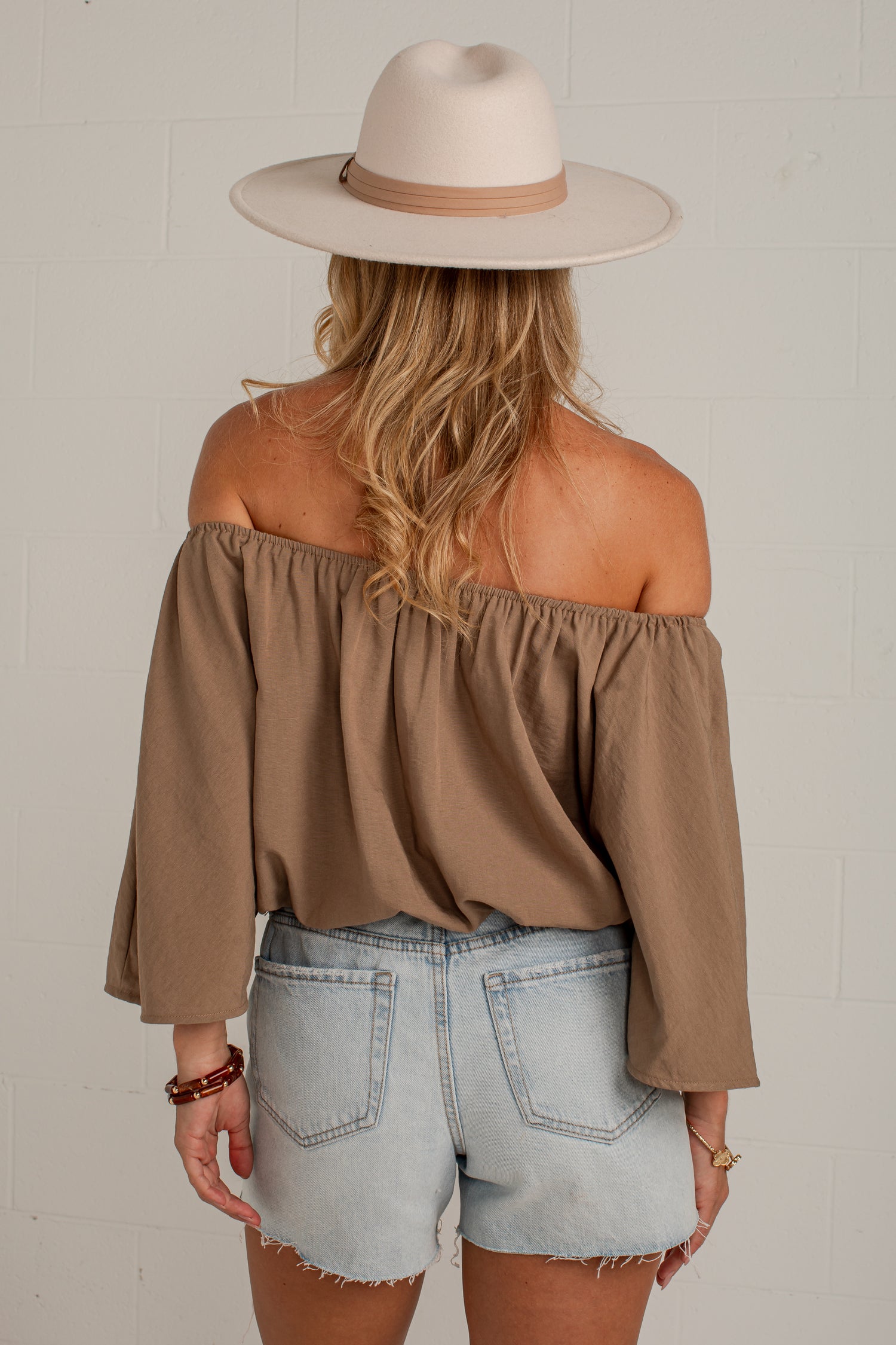 A woman shown from the back wearing a taupe brown off-the-shoulder long-sleeve top, light-wash denim shorts, and a beige wide-brim hat against a white brick wall.