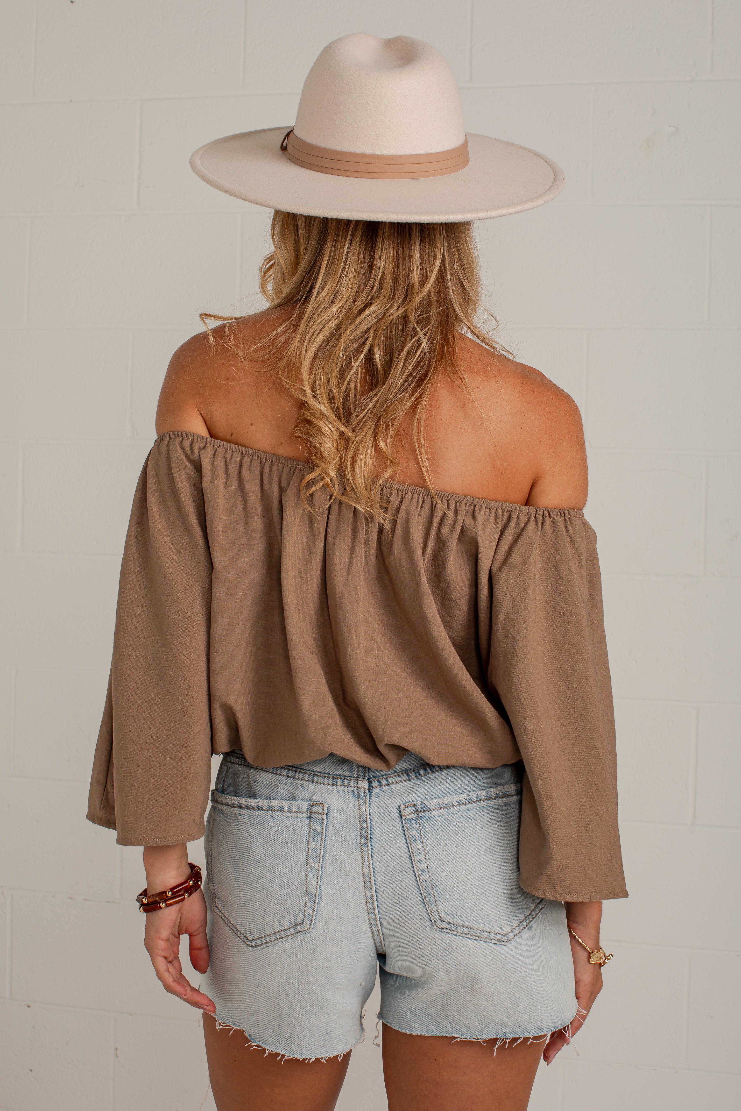 A woman shown from the back wearing a taupe brown off-the-shoulder long-sleeve top, light-wash denim shorts, and a beige wide-brim hat against a white brick wall.