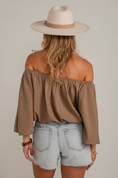 A woman shown from the back wearing a taupe brown off-the-shoulder long-sleeve top, light-wash denim shorts, and a beige wide-brim hat against a white brick wall.