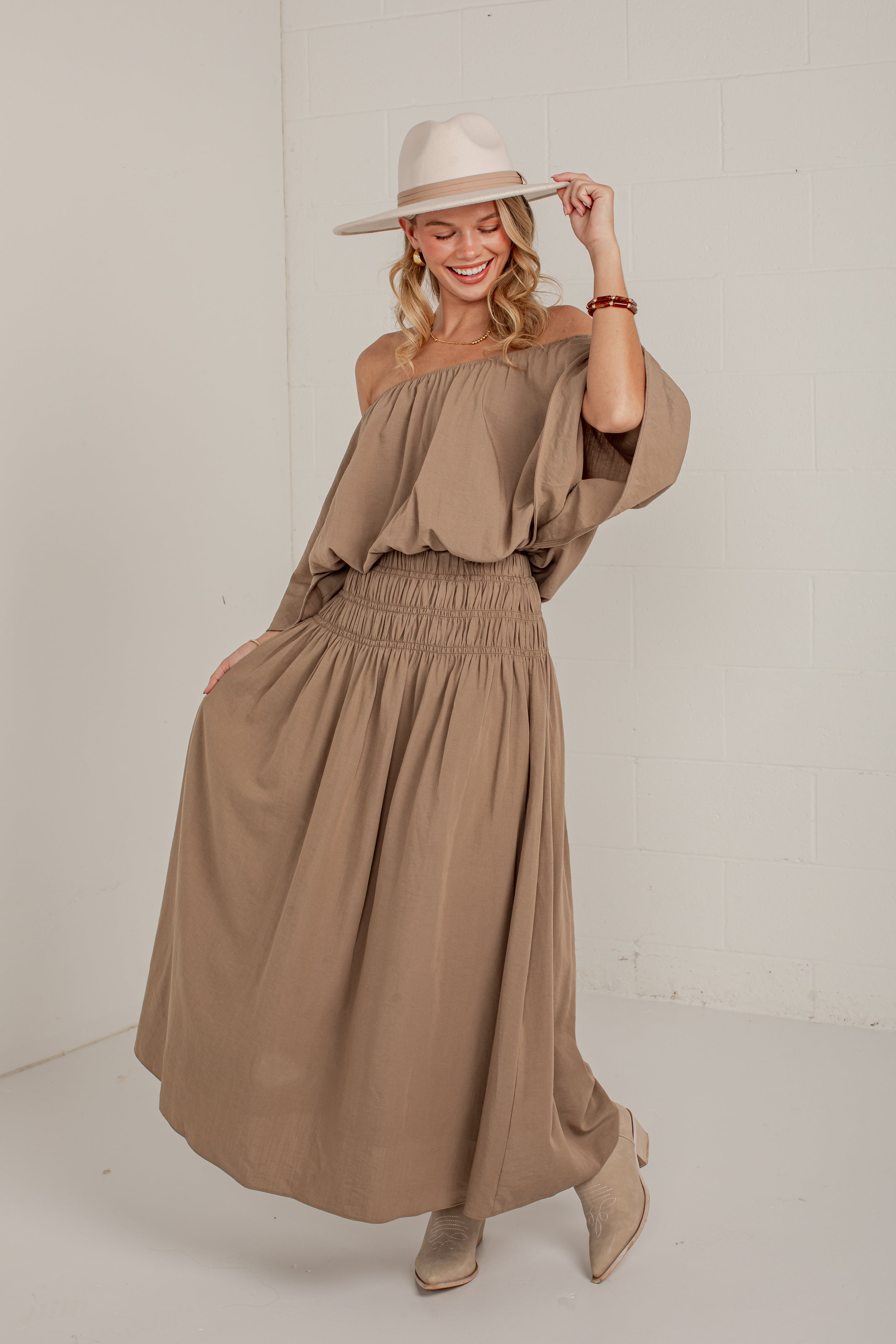 A woman modeling a taupe brown off-the-shoulder maxi dress with a gathered elastic waist, paired with a cream wide-brim hat and beige ankle boots, standing against a white brick wall.