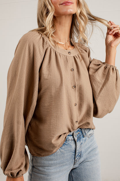 A woman wearing a taupe brown long-sleeve blouse with a gathered neckline and button-front detail, paired with light-wash blue jeans against a white paneled wall.