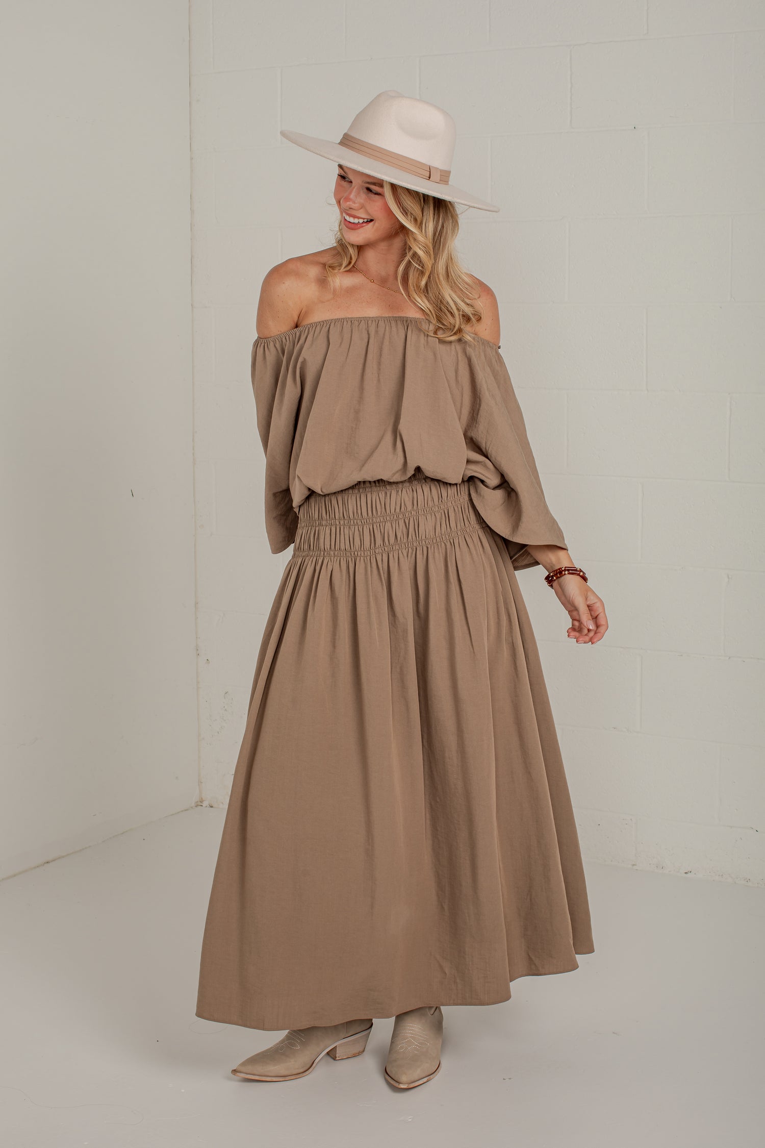 A woman modeling a two-piece taupe off-the-shoulder dress set with a smocked waist, paired with a wide-brimmed cream hat and beige boots, in a bright studio setting.
