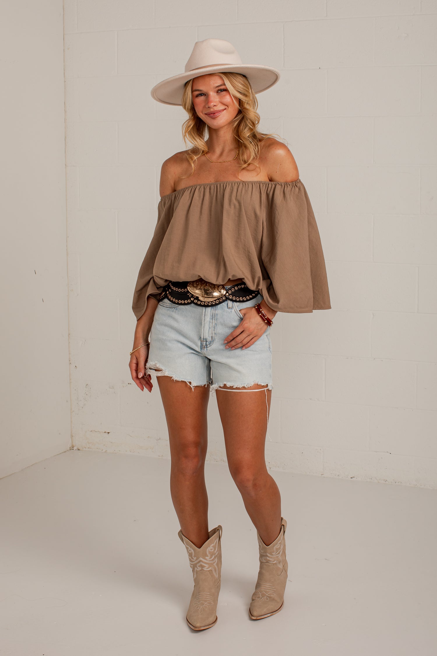 A woman modeling a brown off-the-shoulder blouse with dramatic bell sleeves, paired with light-wash denim shorts, a black belt with gold accents, a beige wide-brim hat, and beige cowboy boots, standing against a white brick wall.