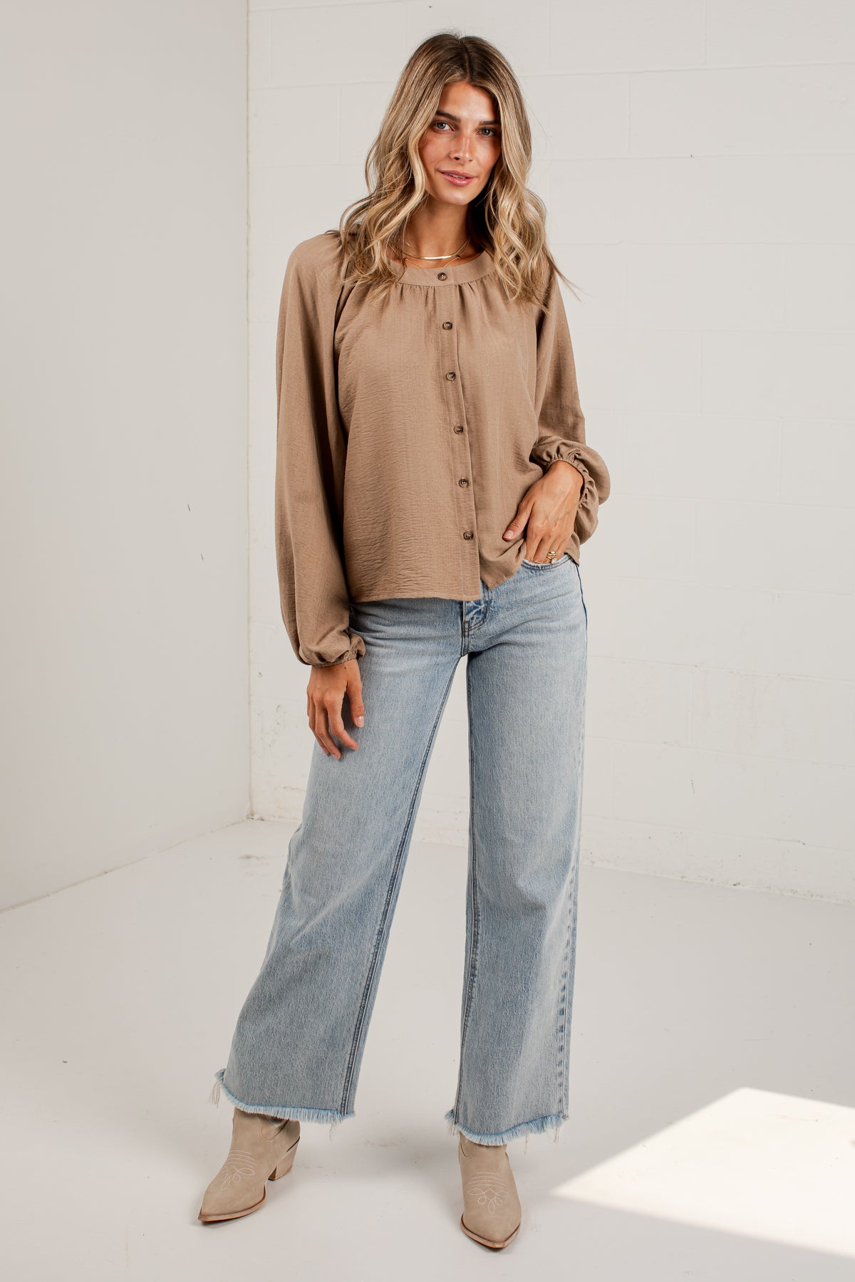 A woman modeling a light brown long-sleeve button-up blouse with ruffled cuffs, paired with light-wash wide-leg jeans and beige ankle boots, standing in a bright white studio.