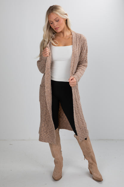 Woman wearing a long beige cardigan, white top, black leggings, and tan knee-high boots on a white background