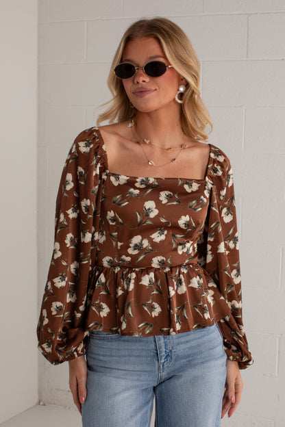 A woman modeling a brown long-sleeve blouse with a white and beige floral pattern, square neckline, and puff sleeves, paired with light blue jeans against a white brick wall.