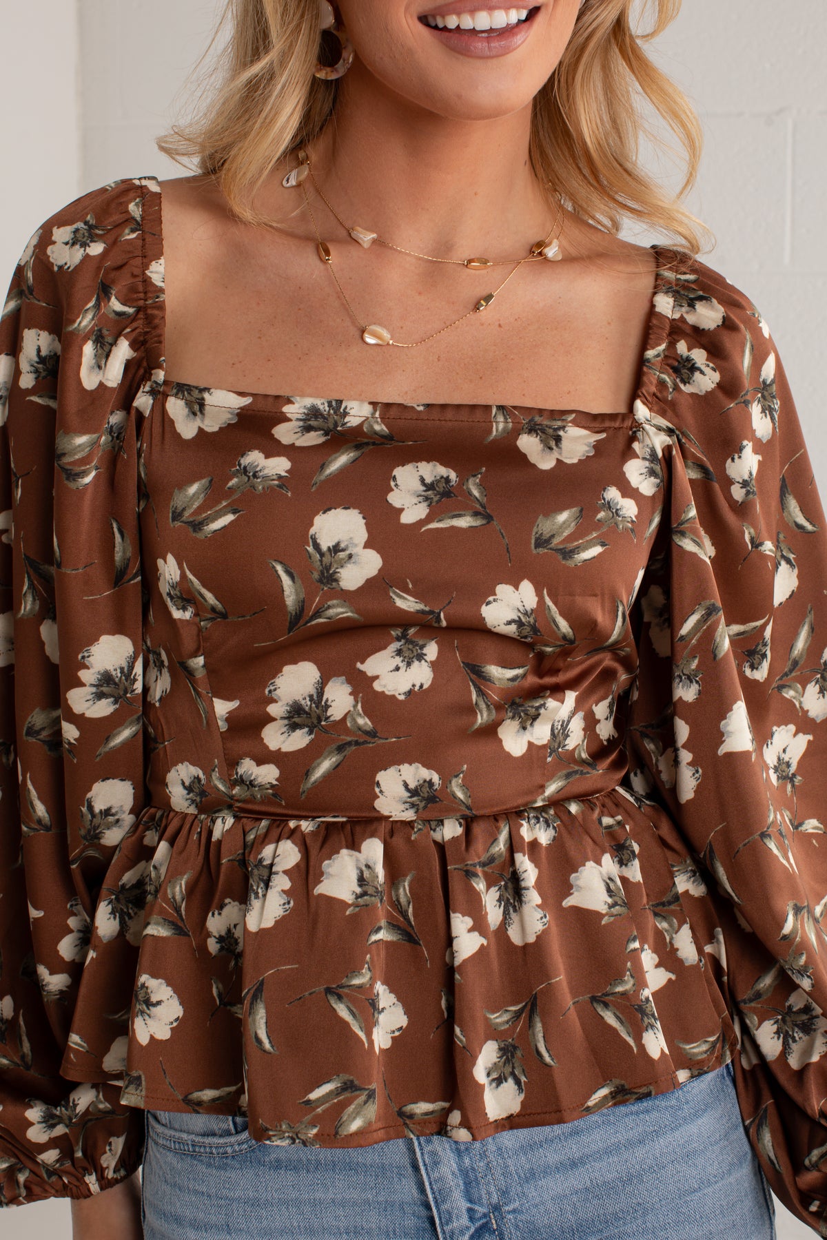 A woman wearing a brown satin-like blouse with a white and beige floral print, featuring a square neckline, puff sleeves, and a gathered peplum hem, paired with blue jeans.
