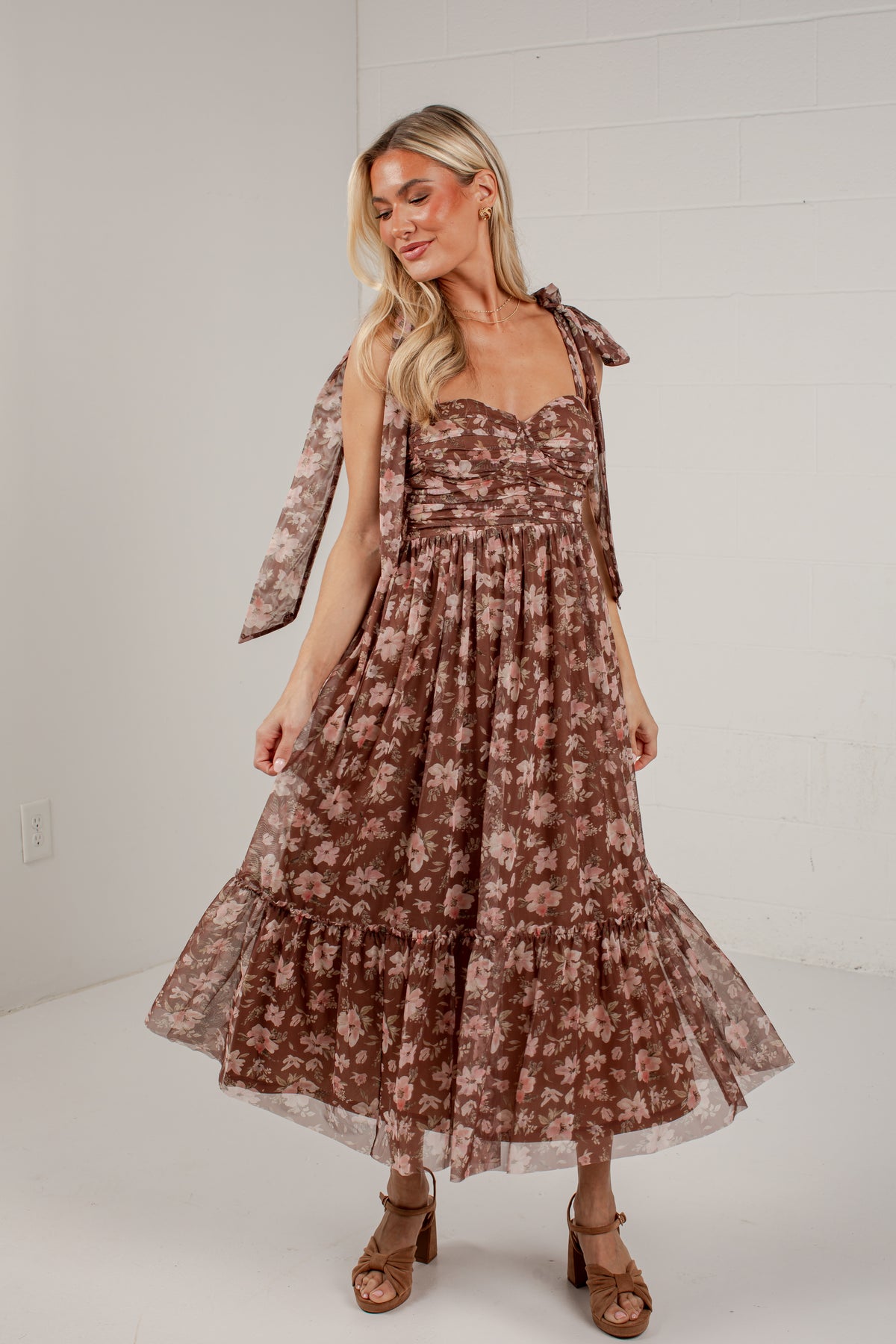 A woman modeling a brown tiered midi dress with a beige floral pattern, featuring a sweetheart neckline, thin shoulder straps with bow details, and a flared skirt, paired with brown heeled sandals against a white brick wall background.