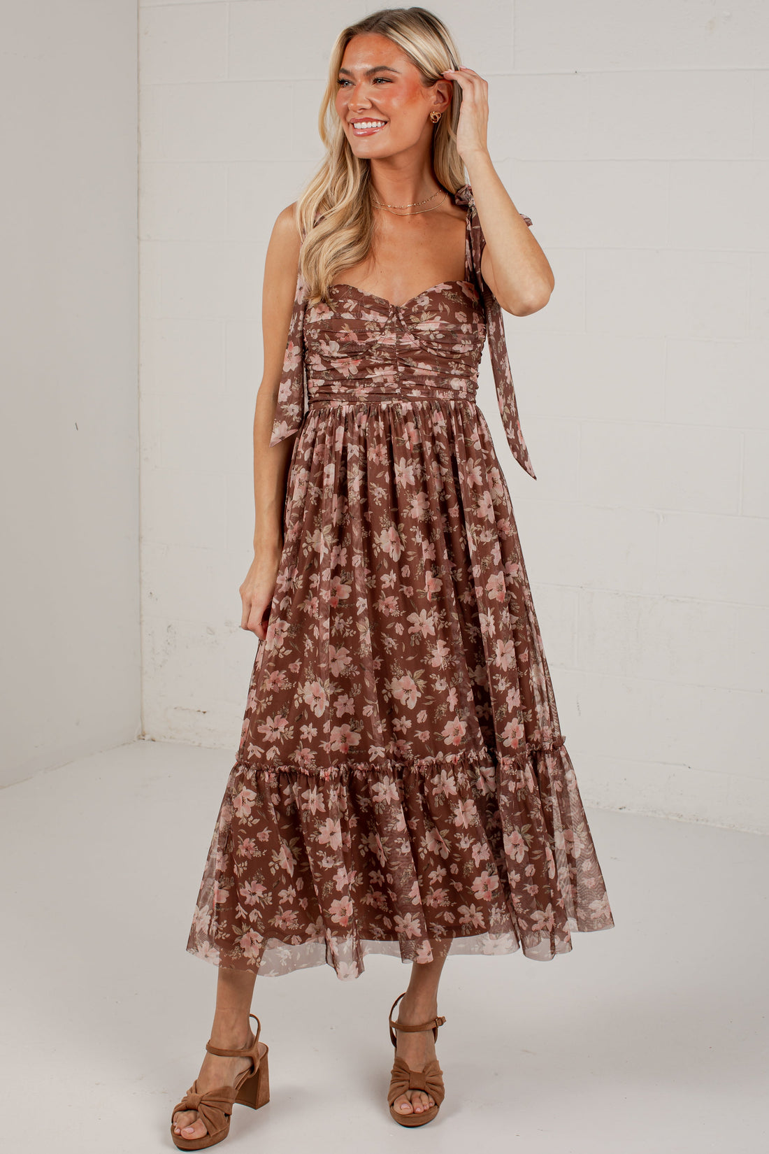 A woman modeling a brown and beige floral tiered midi dress with tie straps, paired with brown heeled sandals, in a bright white studio setting.