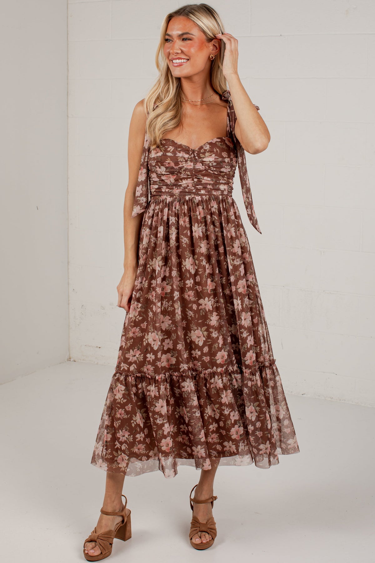 A woman modeling a brown and beige floral tiered midi dress with tie straps, paired with brown heeled sandals, in a bright white studio setting.