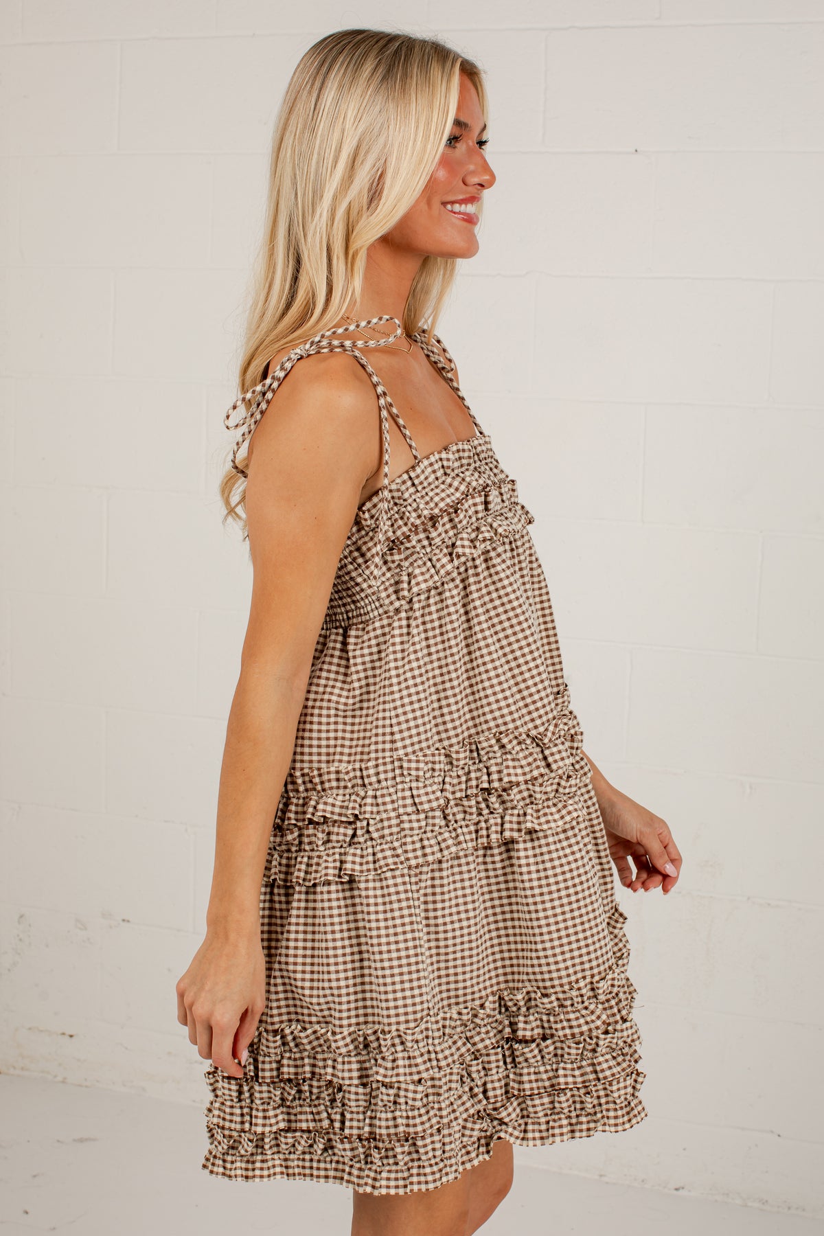 A woman wearing a brown and beige gingham tiered dress with ruffled straps, shown from a side profile against a white brick wall.