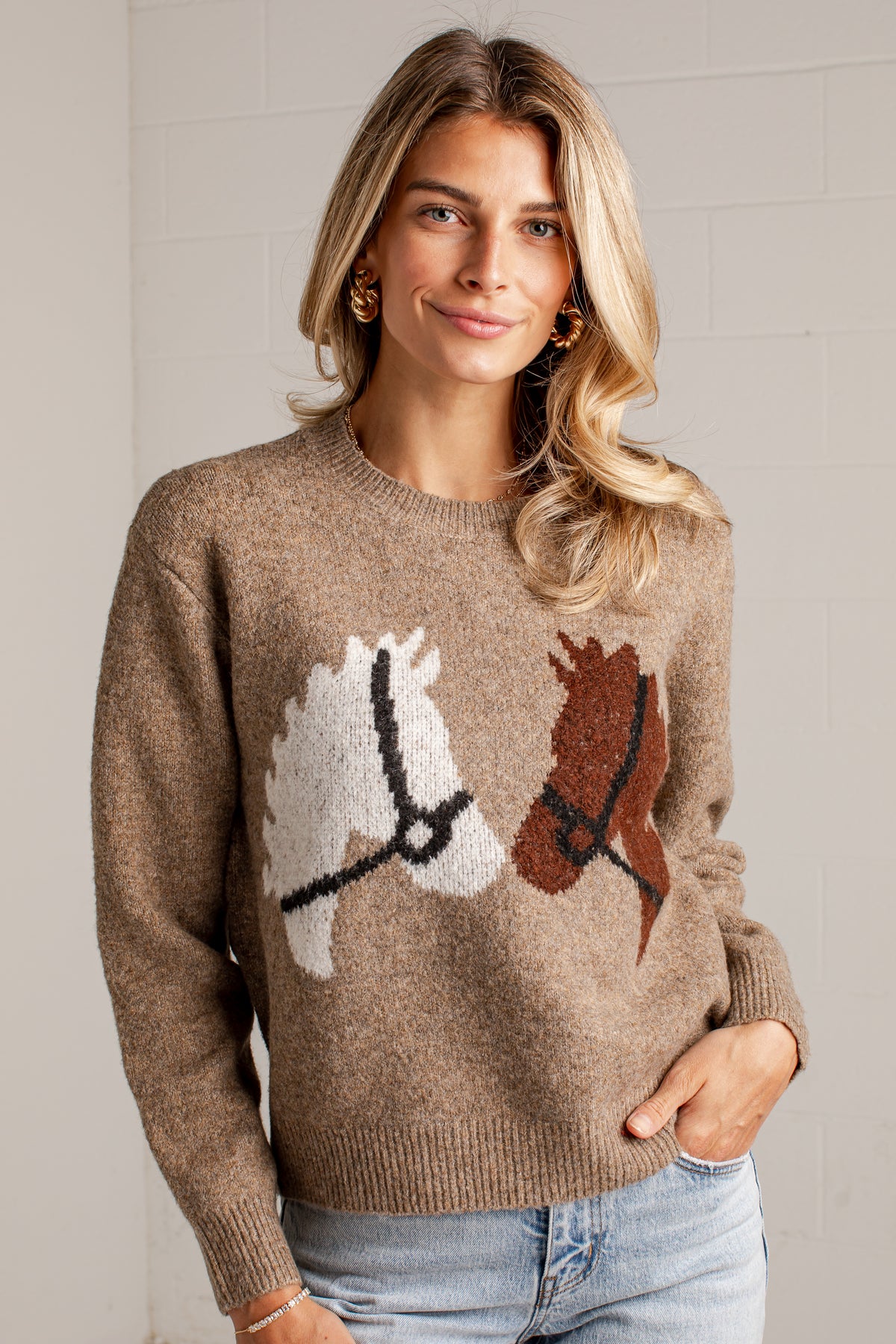 A woman wearing a brown knit sweater with a large horse motif on the front, paired with light blue jeans, standing against a light gray brick wall.