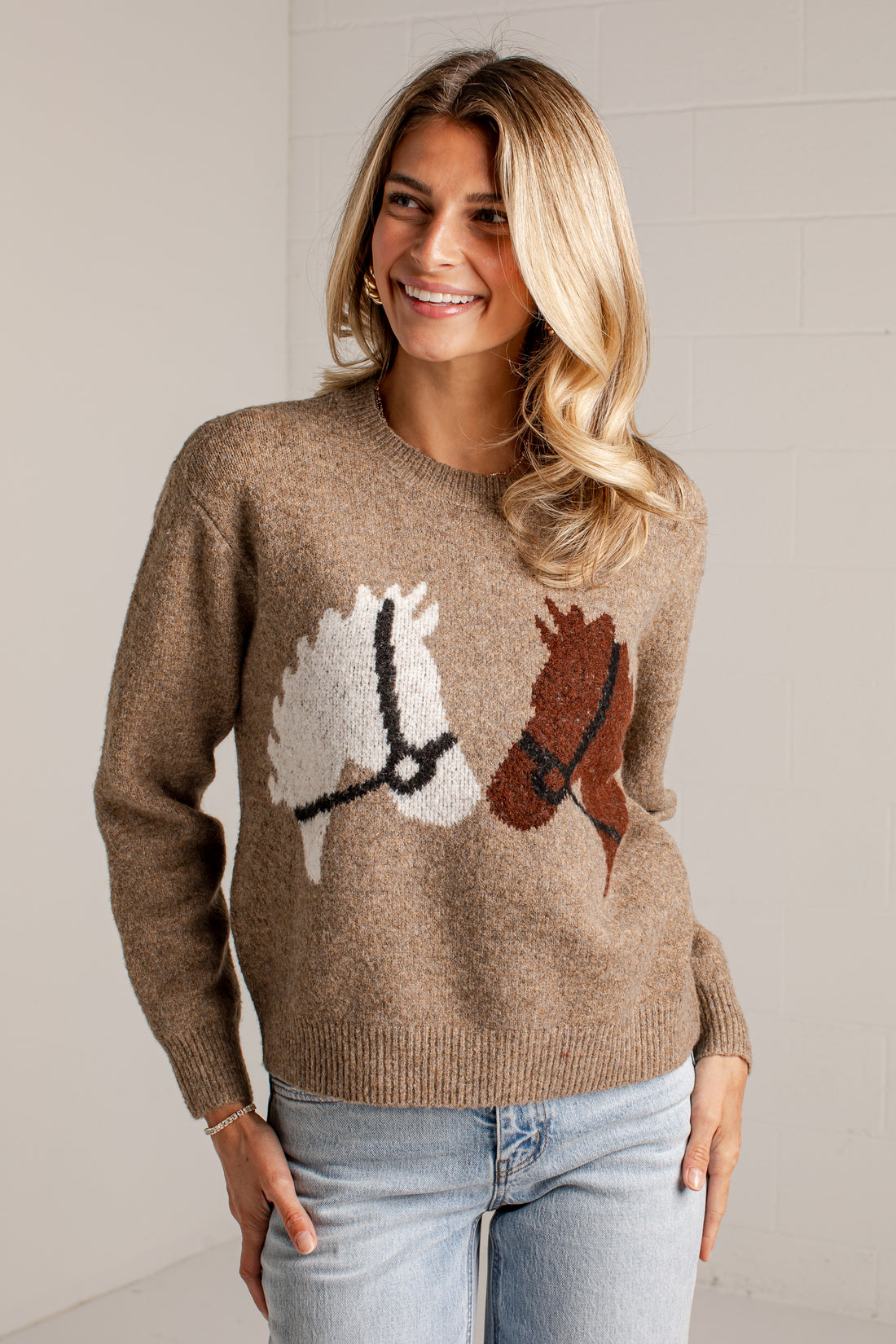 A woman wearing a brown knit sweater with a horse motif and light-wash blue jeans, standing against a light gray wall.