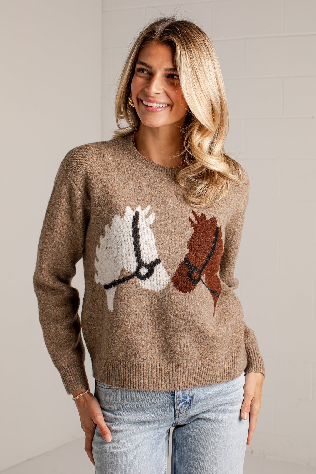 A woman wearing a brown knit sweater with a horse motif and light-wash blue jeans, standing against a light gray wall.