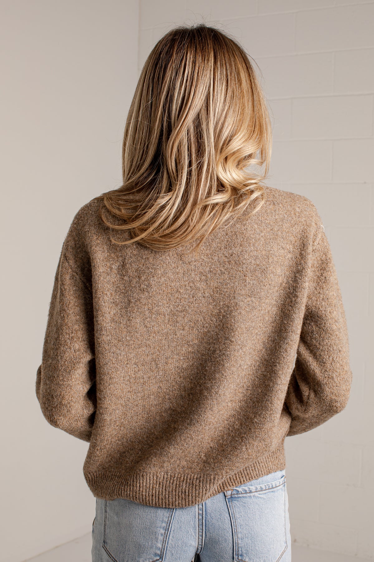 A woman viewed from behind wearing a soft, fuzzy knit sweater in a warm camel brown color, paired with light blue jeans, against a neutral off-white brick wall.