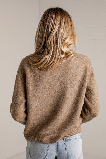 A woman viewed from behind wearing a soft, fuzzy knit sweater in a warm camel brown color, paired with light blue jeans, against a neutral off-white brick wall.
