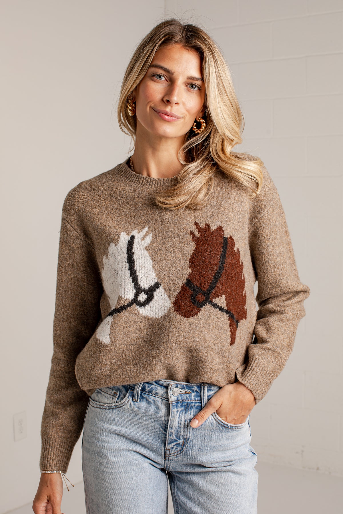 A woman wearing a brown knit sweater with a horse motif and light-wash blue jeans, standing against a light gray wall.