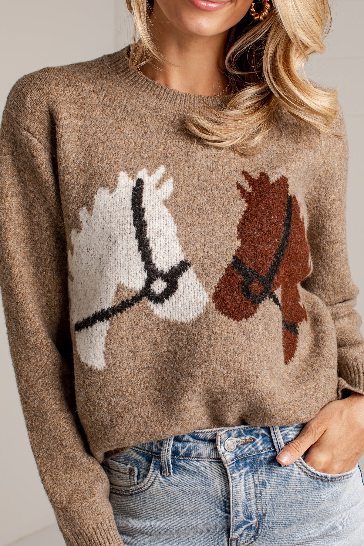 A woman wearing a brown knit sweater with a large horse motif across the chest, paired with light blue jeans. The sweater is a heathered medium brown color with one white horse and one brown horse design, both detailed with black harnesses.