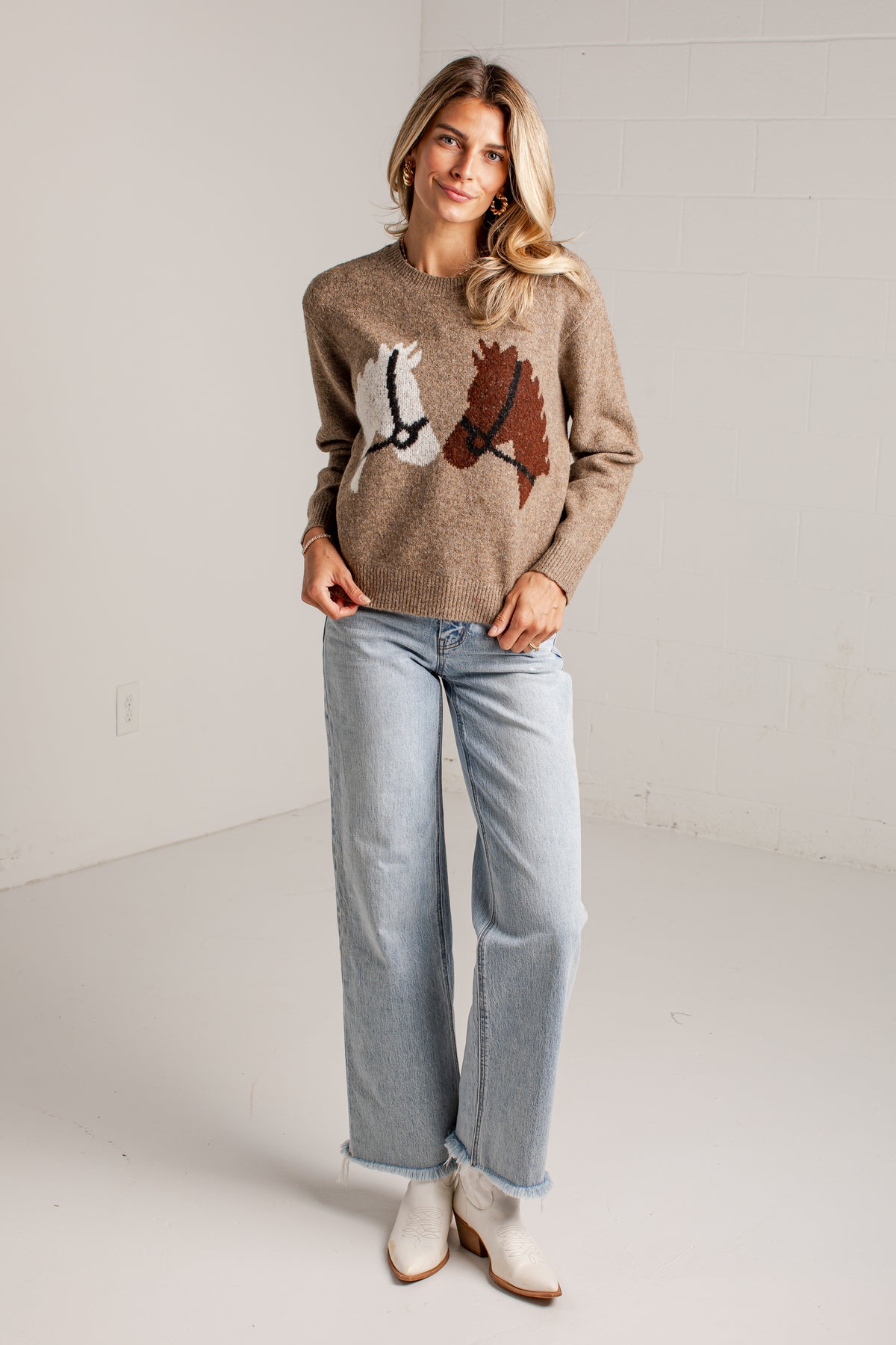 A woman modeling a taupe knit sweater with a horse graphic on the front, paired with light-wash wide-leg jeans and white ankle boots, standing in a bright studio setting.