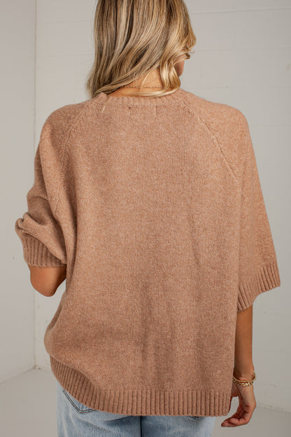 A woman viewed from behind wearing a loose, heathered tan knit sweater with elbow-length raglan sleeves and ribbed cuffs, hem, and neckline, paired with light blue jeans against a white wall.