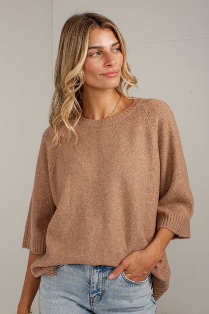 A woman wearing a soft, heathered taupe-brown knit sweater with elbow-length sleeves and a relaxed fit, paired with light-wash blue jeans, standing against a light gray wall.