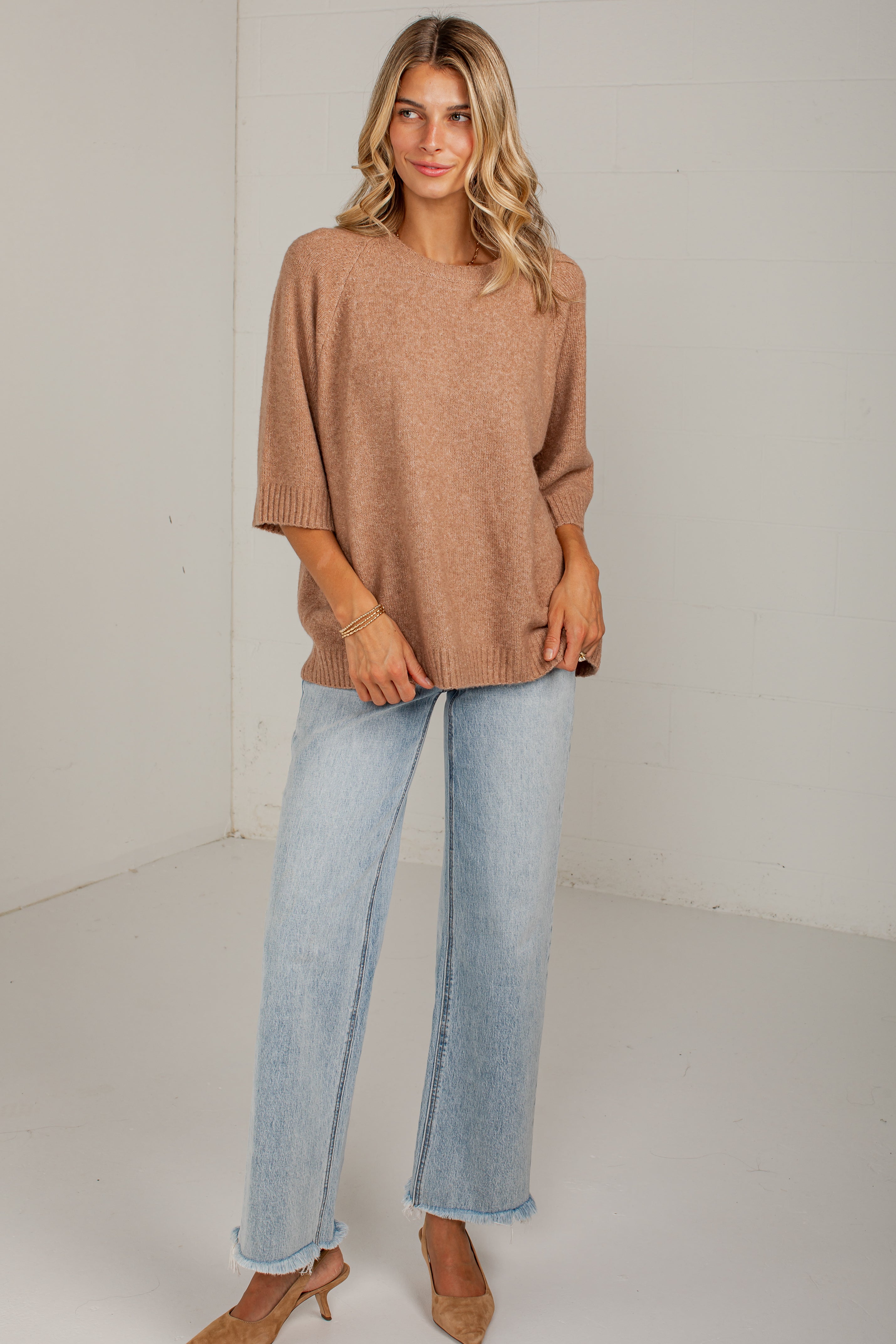 A woman modeling a loose, oversized knit sweater in a warm taupe-brown color, paired with light-wash wide-leg jeans and tan heels, standing against a white studio background.
