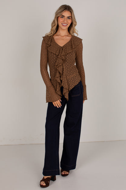 A woman modeling a brown textured knit ruffle cardigan with long bell sleeves, paired with dark wide-leg jeans and brown sandals, against a plain light gray studio background.