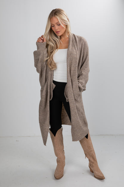 Woman wearing a long beige cardigan, white top, black leggings, and tan knee-high boots on a white background