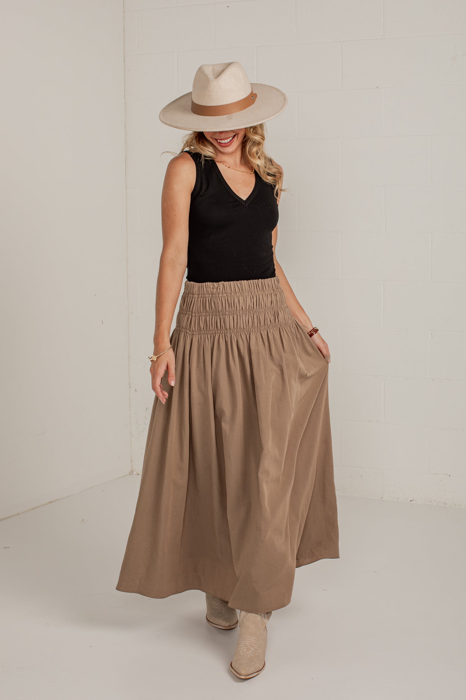 A woman modeling a long, pleated taupe-brown maxi skirt with side pockets, paired with a black sleeveless top, a beige wide-brim fedora hat, and beige ankle boots, standing against a white studio wall.
