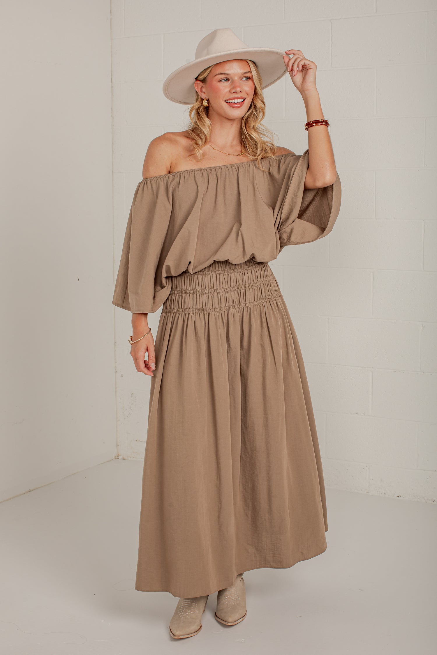 A woman modeling a two-piece taupe off-the-shoulder top and matching smocked maxi skirt, styled with a wide-brimmed beige hat and beige ankle boots against a white studio wall.