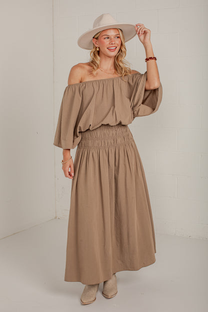 A woman modeling a two-piece taupe off-the-shoulder top and matching smocked maxi skirt, styled with a wide-brimmed beige hat and beige ankle boots against a white studio wall.