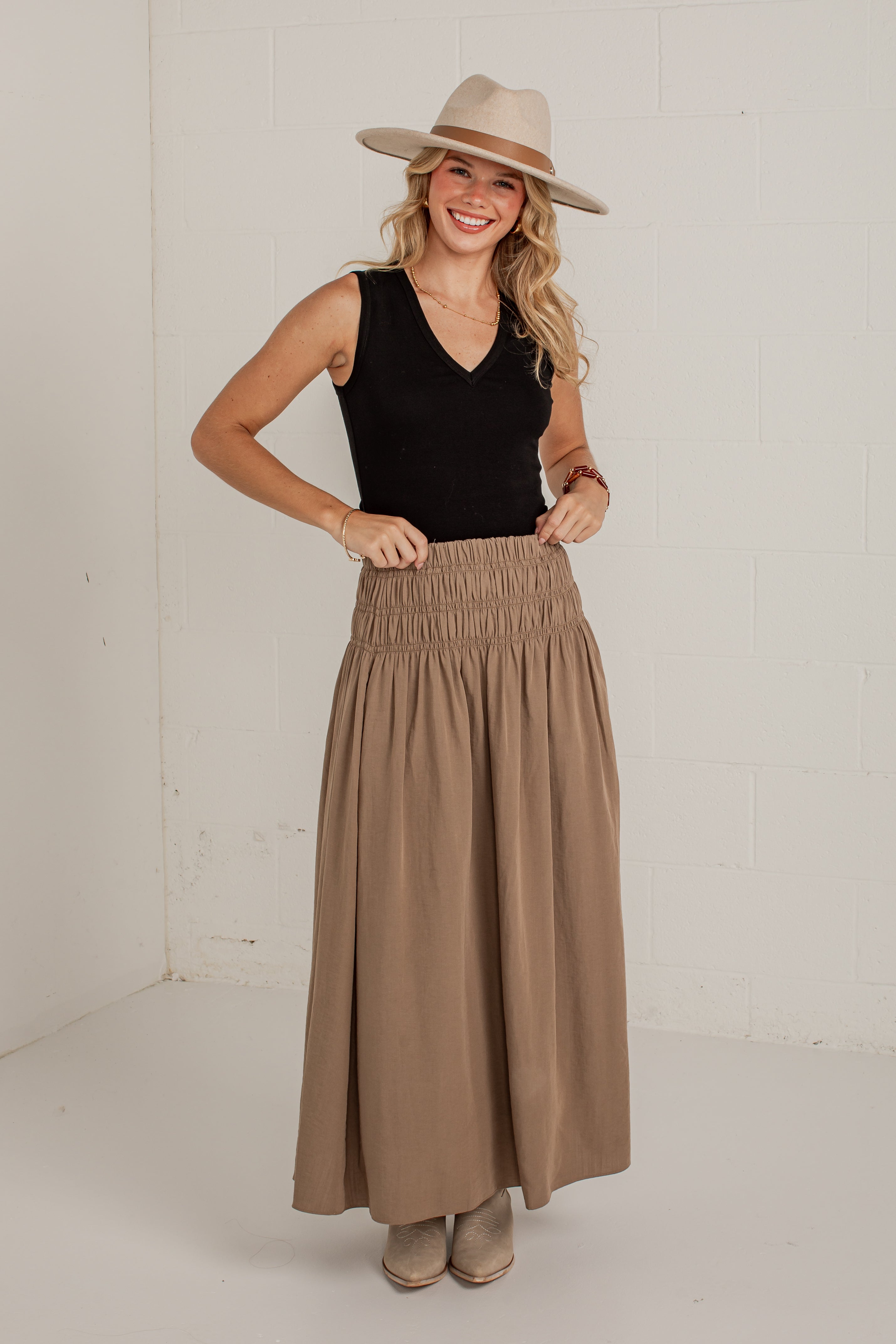 A woman modeling a high-waisted, smocked wide-leg maxi skirt in taupe brown, paired with a black sleeveless top, a beige wide-brim hat, and beige boots, standing against a white brick wall.